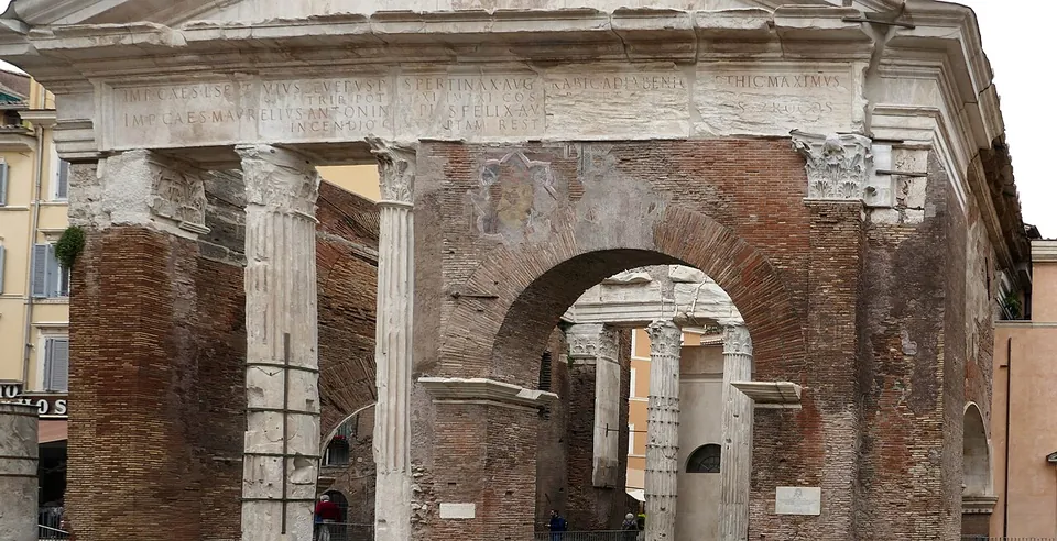 The front of a ruined gateway, Corinthian columns and an inscribed architrave under a pediment, with a large brick arch filling the space where two columns once stood.