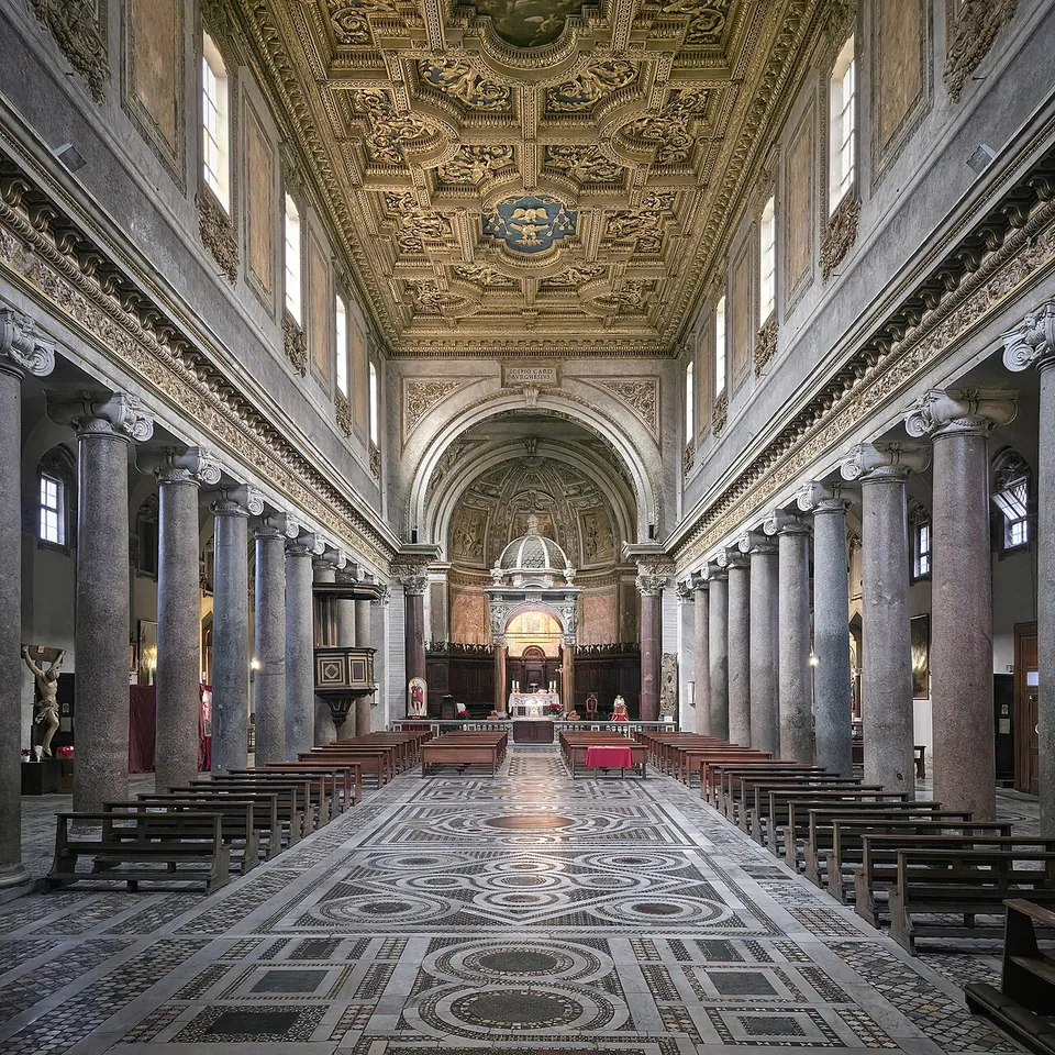 The interior of a long church divided by rows of grey granite columns, with a floor of inlaid marble in circles and bands, a gilded coffered ceiling, and a canopy over the altar at the far end in front of a painted apse.