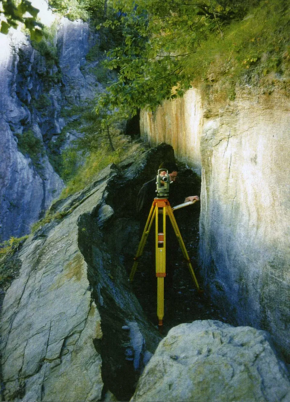 A channel cut along a sheer rock face high in a gorge, with a wall of rock left standing on the valley side and a surveyor's tripod set up in the channel.