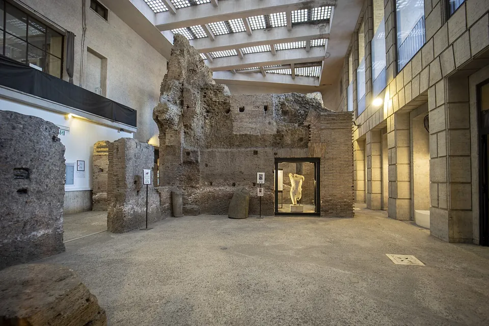 A wide underground hall lit by a skylit modern ceiling, where tall broken walls and piers of brick and stone rise from a concrete floor, with information panels set up between them.