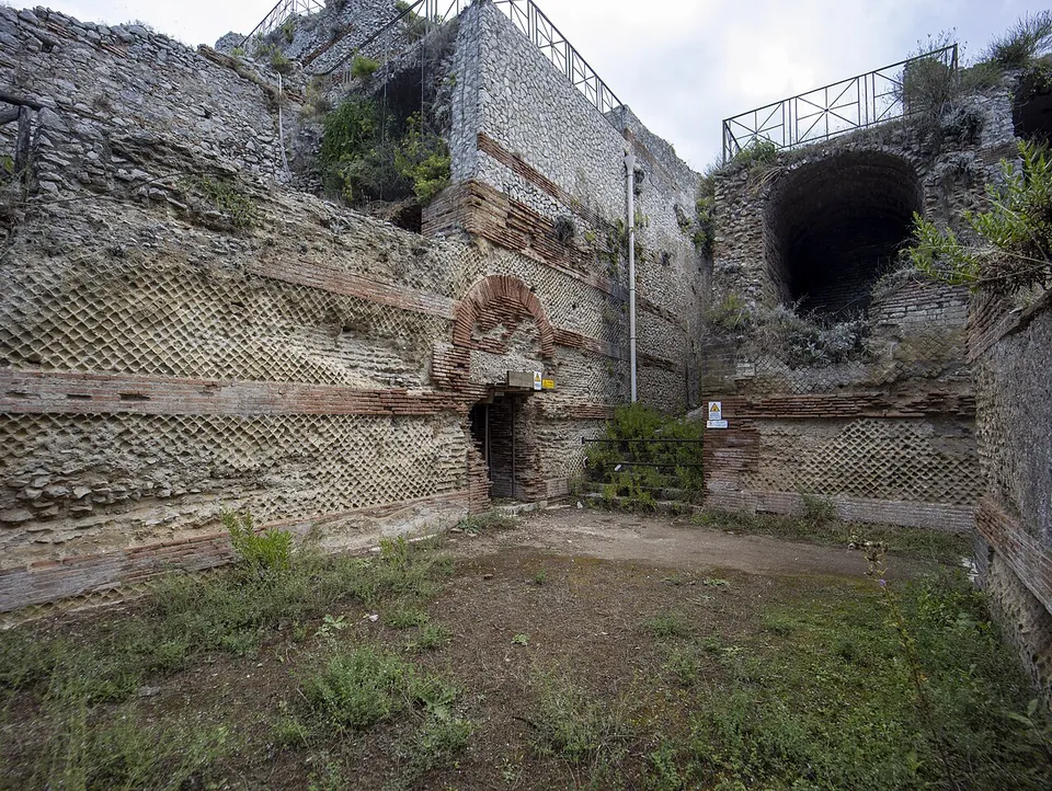 Tall walls faced in a diamond-net pattern of small square stones, banded with courses of red brick, around an open room with a grassy floor; a brick arch frames a doorway, and above it a large vaulted opening with a railing on top.