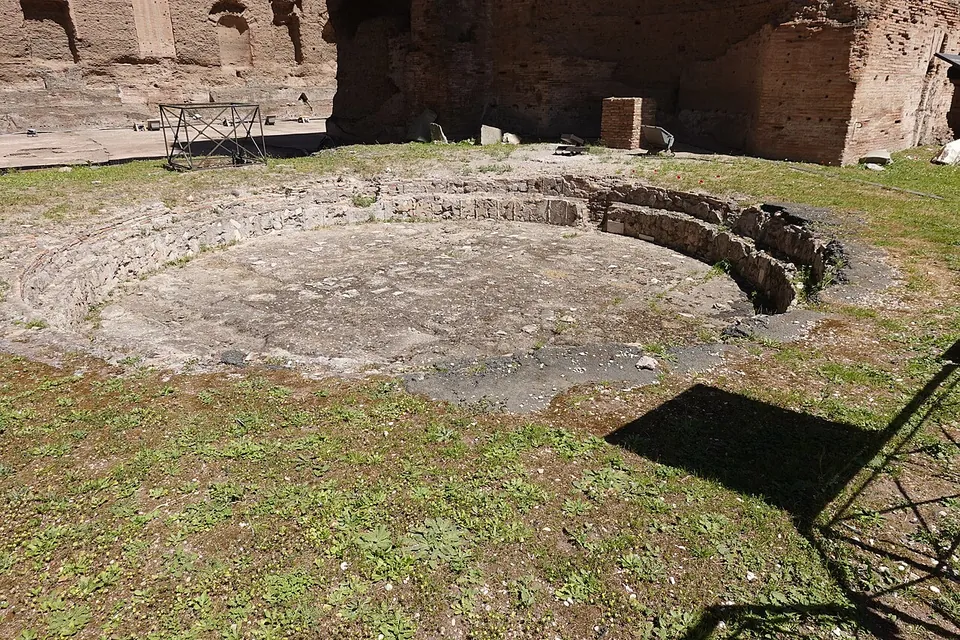 A shallow circular basin with stepped sides set into the ground among grass, with ruined brick walls behind it.