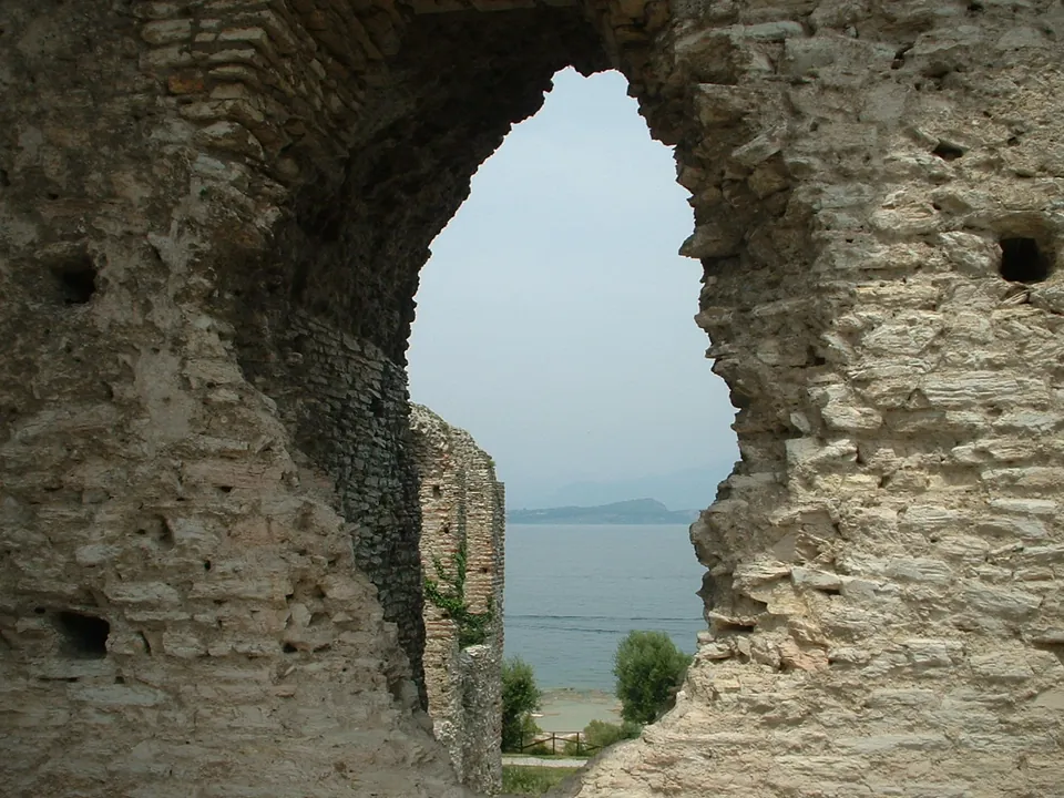 Close view of a broken arched opening in a thick wall of small stones laid in courses, with small square holes in the masonry and the lake and a beach visible through the gap.