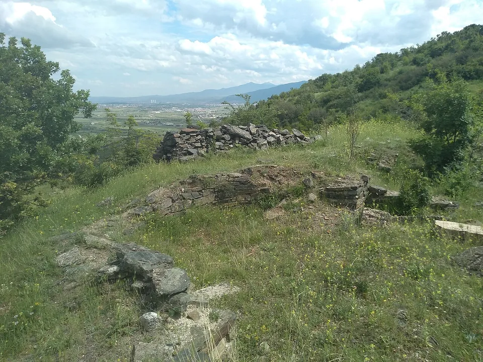 Low courses of flat stones outlining walls in the grass of a hilltop, with bushes around and a wide plain with a city and mountains in the distance.