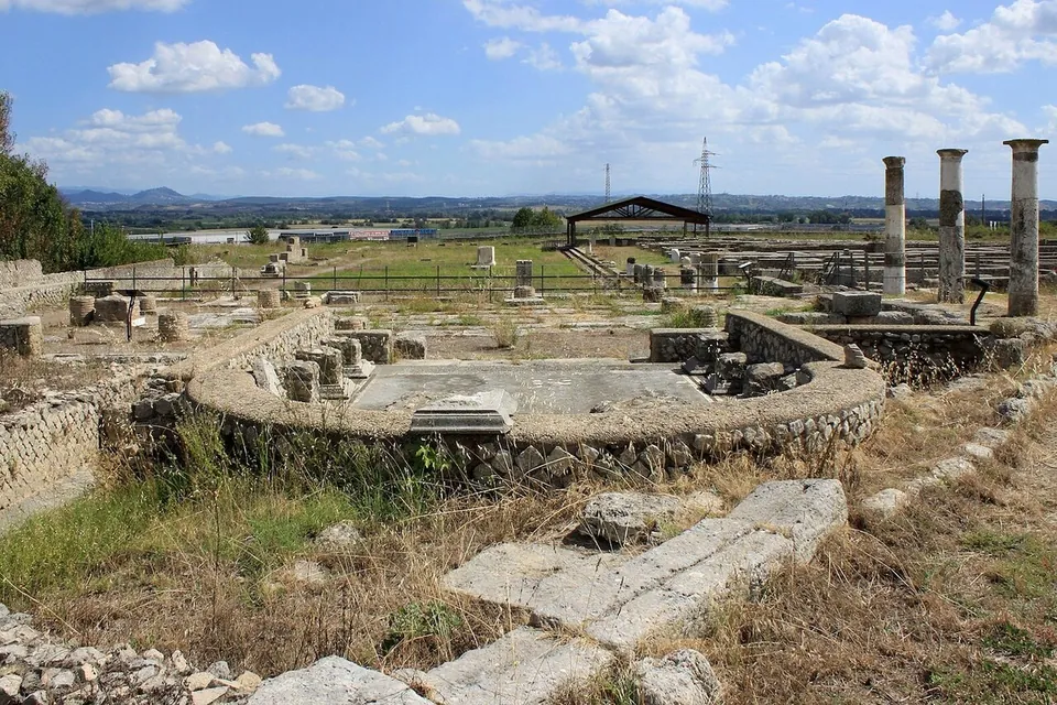 In the foreground a curved wall of rubble masonry encloses a paved floor; beyond it three tall columns, low walls and a modern canopy roof stand on a flat plain, with hills along the horizon.
