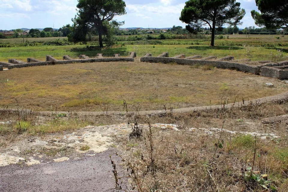 A grassy, nearly round arena ringed by a low wall with short stubs of masonry along its top, and umbrella pines and fields beyond.