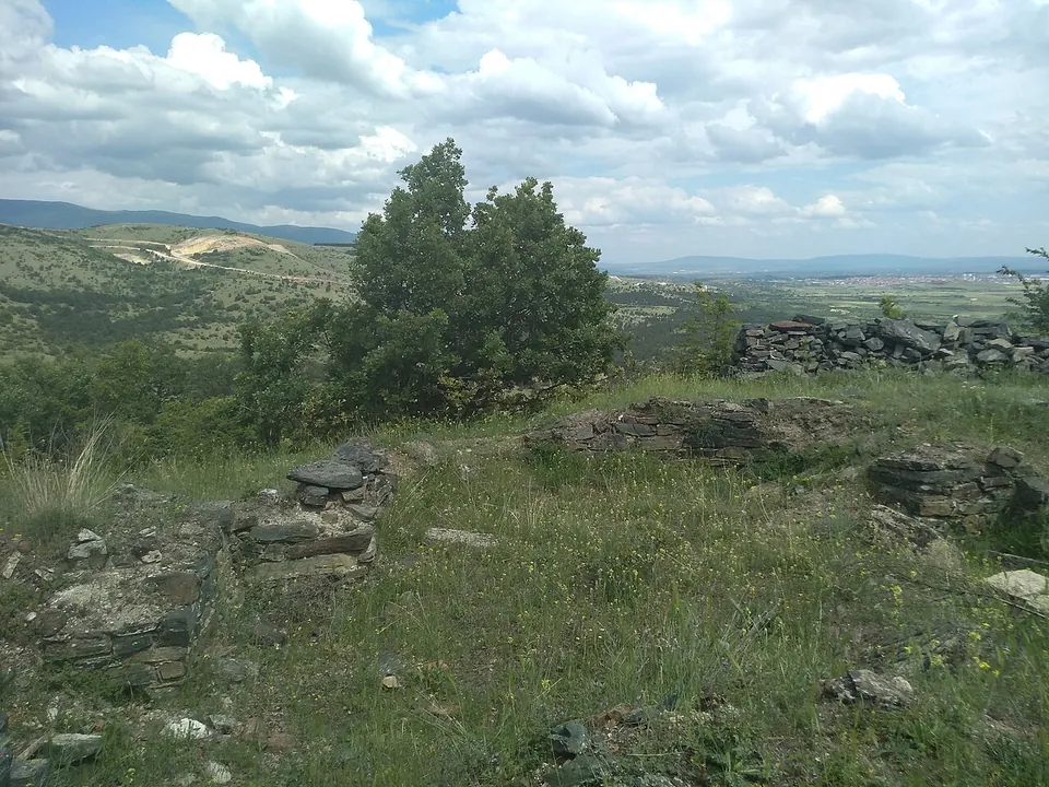 Low stone walls forming the corner of a building on a grassy hilltop, a lone tree behind and hills and a valley stretching away under a cloudy sky.