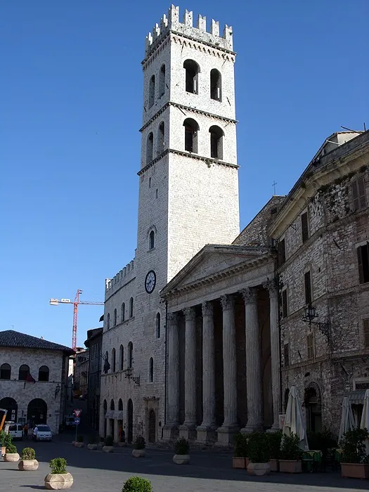 The tall battlemented stone tower of the square rising beside the temple front, its columns and pediment set into a row of later stone buildings along a wide paved square under a clear blue sky.