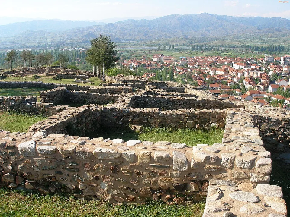 Conserved low walls of rough stone forming a row of rectangular rooms on a grassy hilltop, with the red roofs of a town below and a line of mountains beyond.