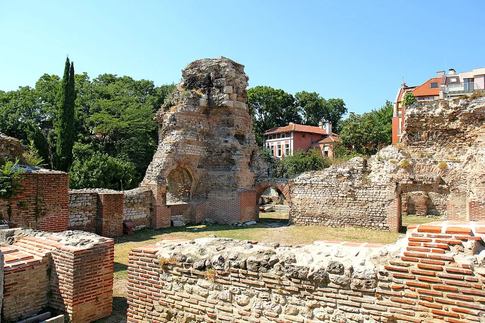 A tall, ragged mass of Roman masonry with a brick arch at its foot rising above low walls of brick and stone, with a grassy floor between them, trees and a red-roofed house behind.