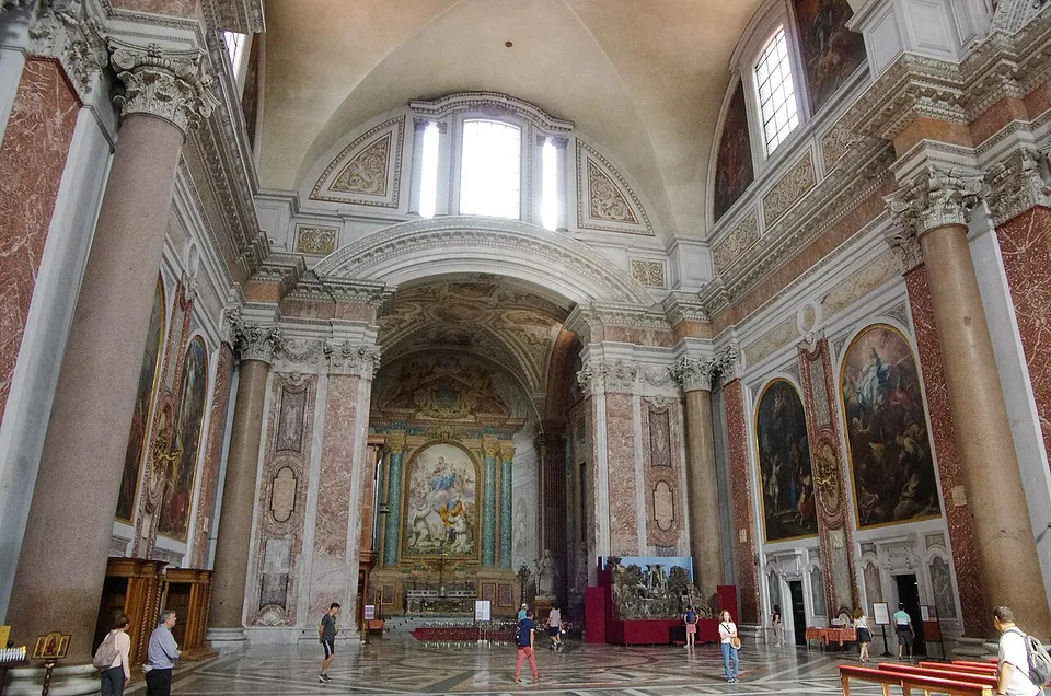 A vast church interior with a patterned marble floor, tall columns with gilded capitals on both sides, large paintings on the walls, and a vaulted ceiling leading to an altar in a lit apse.