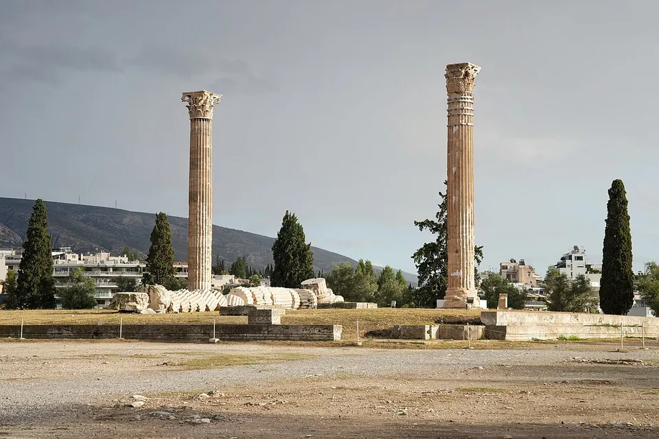 Two lone colossal columns standing apart on a dry open precinct, with the drums of a fallen column lying in a line on the ground between them, cypresses and a hill behind.
