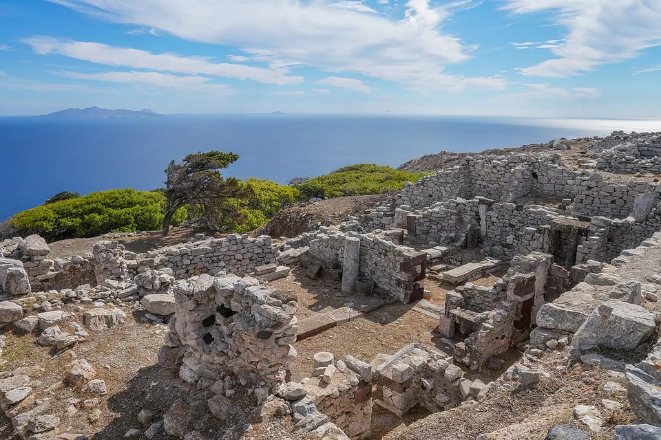 Roofless stone houses with doorways and room walls on a rocky slope, a wind-bent tree and green shrubs beyond, and a deep blue sea with an island on the horizon.