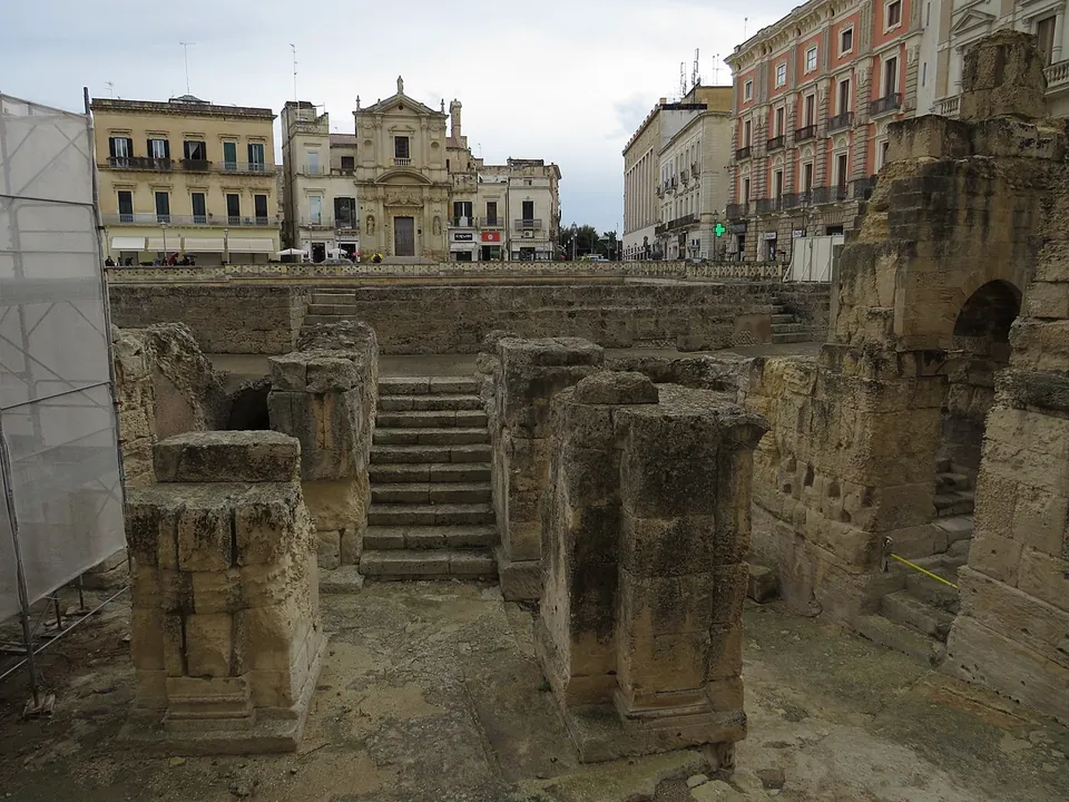 Weathered square stone piers standing in an excavated corridor, a flight of stone steps rising between them towards the square, and a church facade among the buildings beyond, with protective sheeting at the left edge.