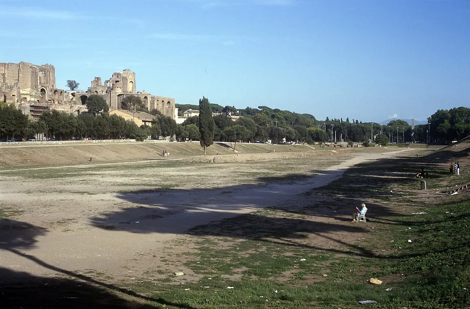 A long, flat valley floor of dry grass and bare earth running into the distance between lines of trees, with tall brick ruins on the left skyline and a single cypress standing near the middle.