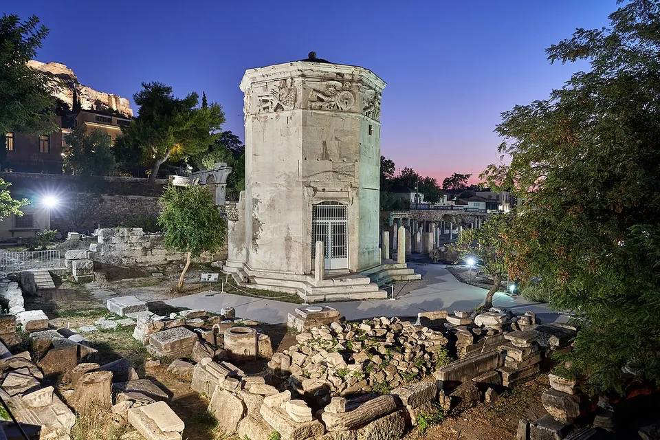 An octagonal marble tower with a band of carved figures below its roof and a small door at its foot, lit at dusk among scattered fallen blocks, with the floodlit Acropolis rock behind.