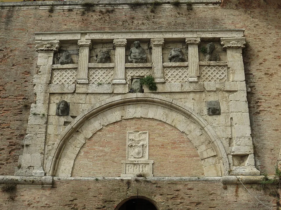 A pale stone gate front set into a tall wall of brick. A wide round arch, now walled up with brick, is framed by pilasters and topped by a balustrade divided by small columns, with weathered sculpted figures between them. Below it a small arched doorway with steps opens in the brick wall.