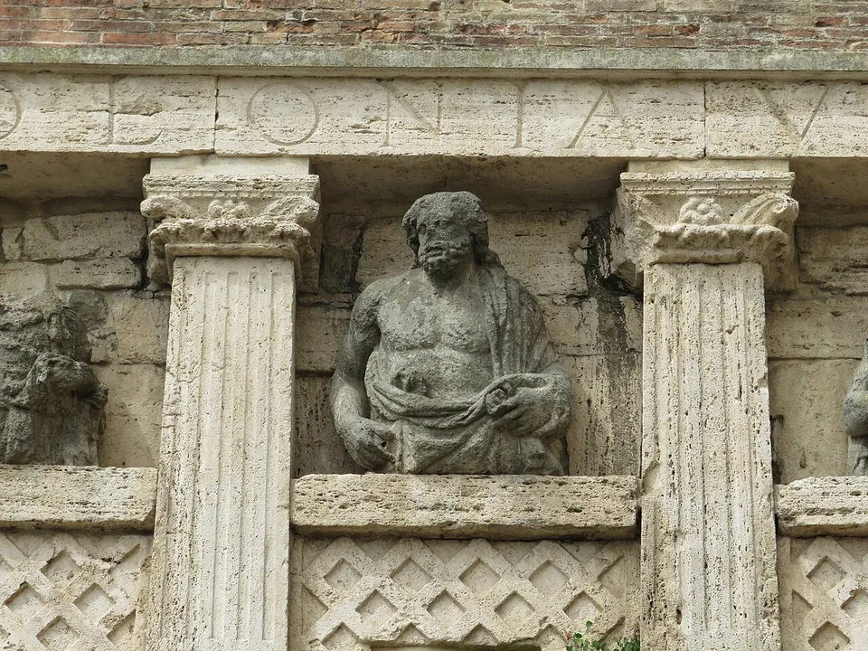 Close-up of a weathered stone half-figure of a bearded, bare-chested man draped in a cloak, set between two fluted pilasters with carved capitals, above a panel of lattice pattern. Large carved letters run along the band at the top.