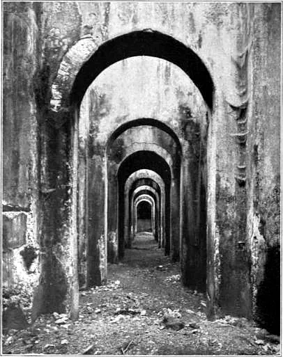 A black-and-white photograph down a long aisle of the cistern, a line of round arches on massive piers receding into darkness over a rubble-strewn floor.
