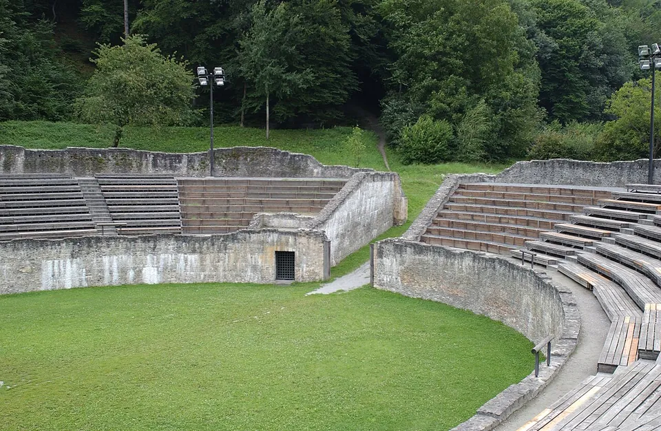 A grassy oval arena ringed by a stone wall, with rows of seating rising in blocks behind it and a small doorway in the wall, set against a wooded slope.
