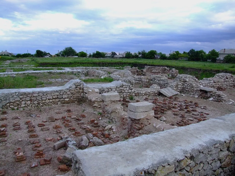 An excavated room enclosed by low stone walls, its floor set with rows of short stacks of red tiles that once held up a heated floor, with more ruins and a village behind.