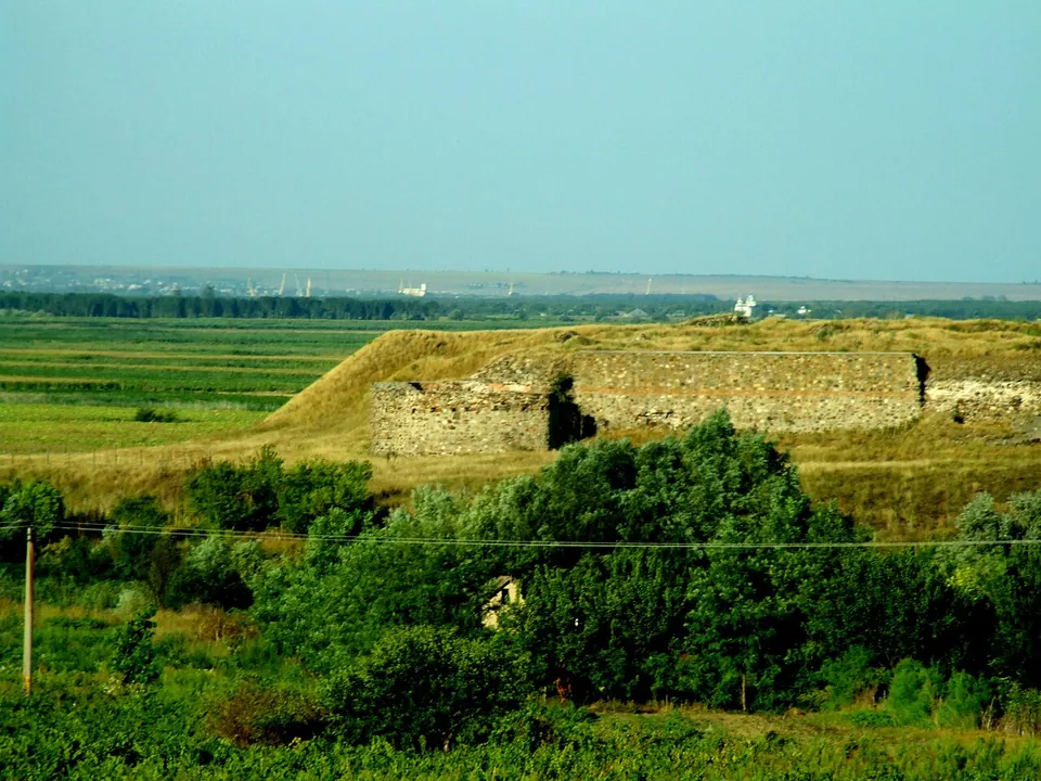 Grey stone walls with rounded towers running along the top of a low grassy rise above green fields and scrub, with the flat plain and a distant town on the horizon.