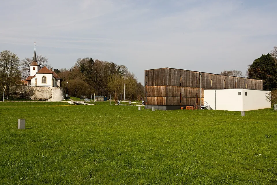A wide lawn with a long, dark timber-clad building and a white annex on the right, and a small white church with a pointed bell tower on a terrace on the left.