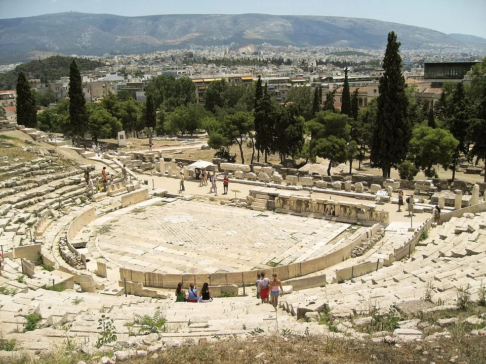 A theatre seen from high in its stone seating, the lower rows curving round a paved semicircular floor, with a low wall of carved reliefs across the front of the stage, cypresses and pines behind and the city spreading to the hills beyond.