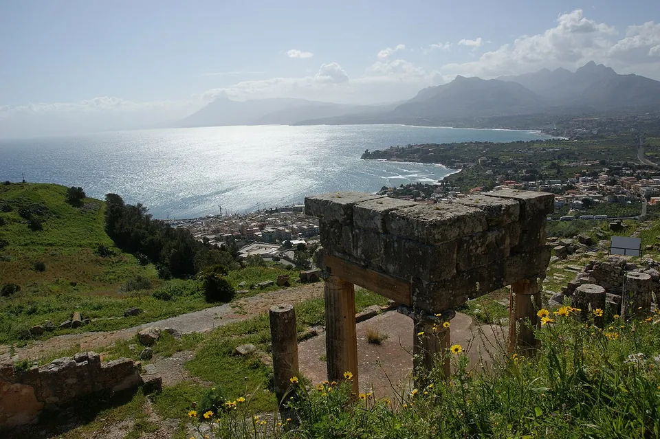A heavy stone entablature block propped on two stone posts and modern wooden supports, standing among ruins on a hillside above a bay, with a town along the shore and a mountain range across the water.