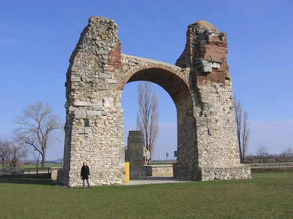 A tall ruined arch of rough grey stone with bands of red brick, standing alone in a flat grassy field under a blue sky, a person at its foot for scale.