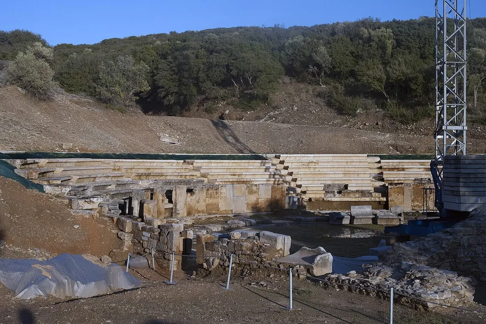 Rows of new and old stone seats behind the ruined stage building, with a steel scaffolding tower at the right and plastic sheeting on the ground.