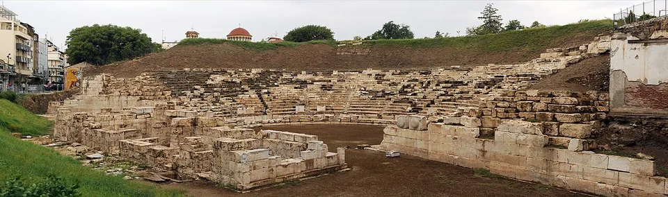 A panorama of a large ancient theatre on a grassy slope, with blocks of marble seating and the stage ruins in front, and city buildings on the left.
