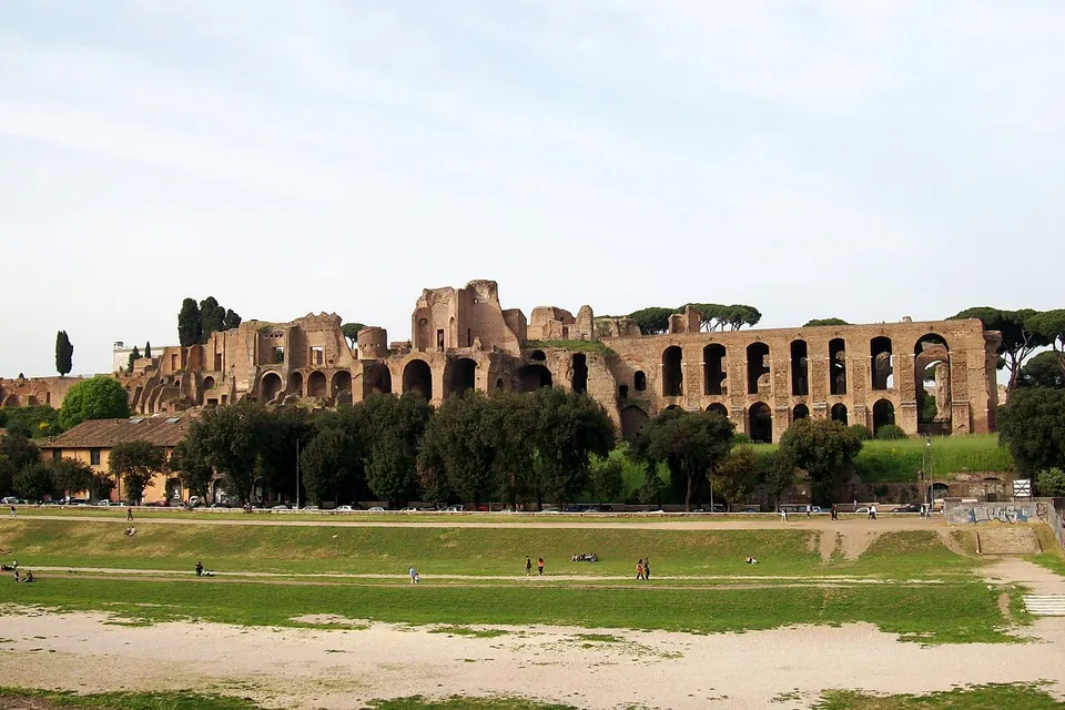 A long ruined brick palace front along the top of a hill, tiers of tall arches and broken walls above a band of trees, with the grassy, sandy floor of the Circus Maximus in the foreground and a few people walking on it.