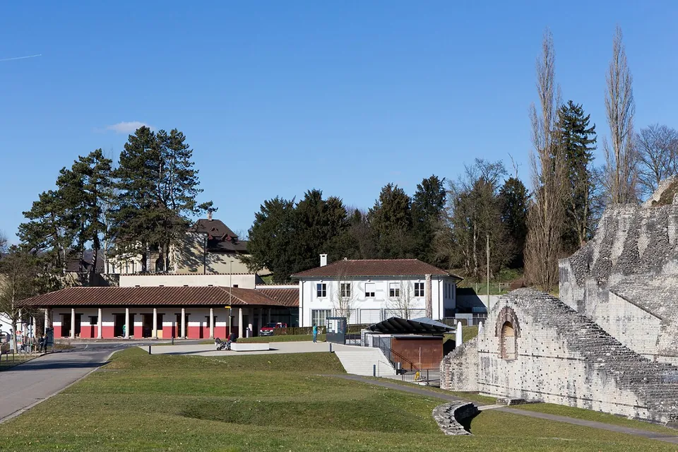 A lawn and paths in front of a long low building with a red-columned arcade and a white house with a hipped roof, with the tall rough stone wall of the theatre rising on the right.