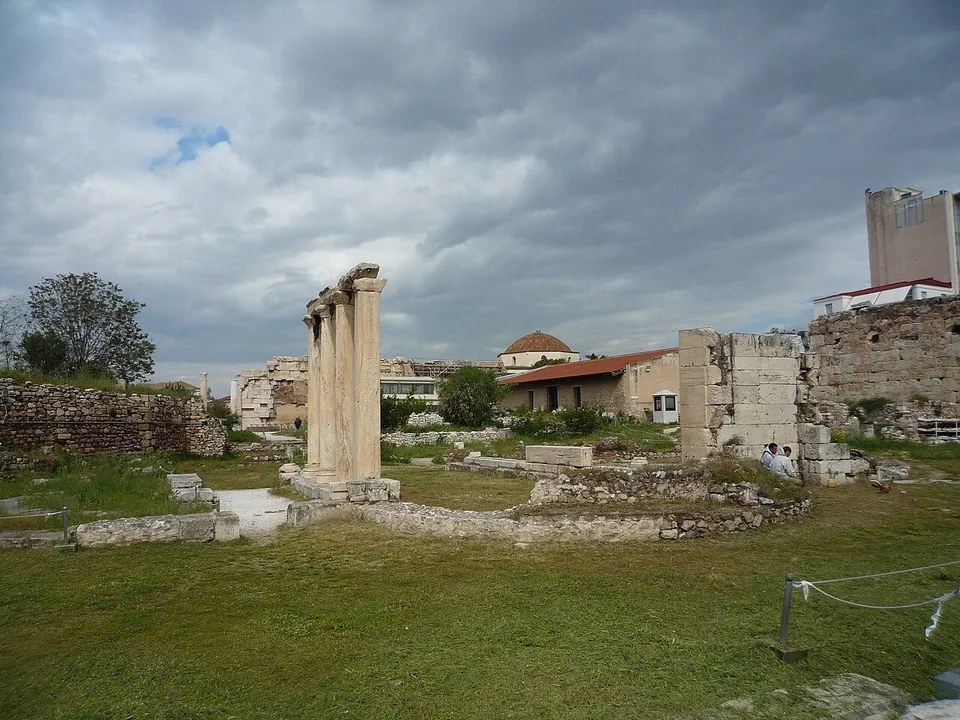 An enclosure of grass and low excavated walls under a stormy sky, with a few columns standing together in the middle distance, a block of masonry on the right and houses beyond.