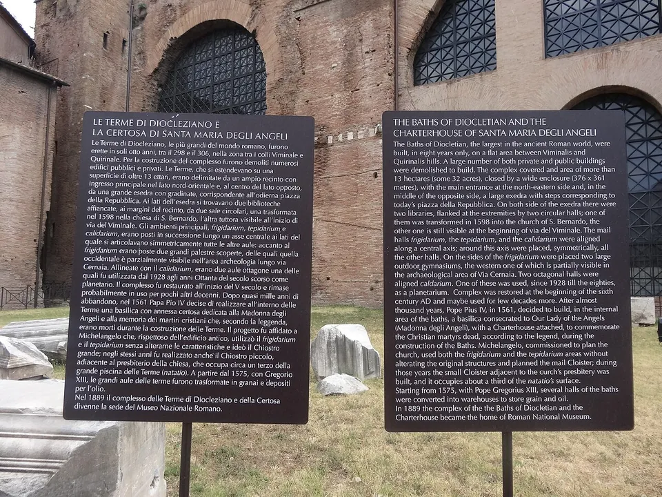 Two dark information boards on posts in front of a brick wall with tall arched windows, one in Italian and one in English, headed "Le Terme di Diocleziano e la Certosa di Santa Maria degli Angeli" and "The Baths of Diocletian and the Charterhouse of Santa Maria degli Angeli".