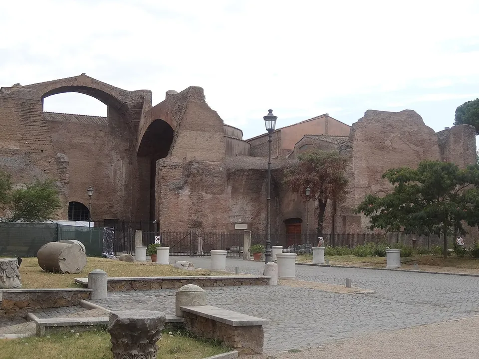 Tall ruined brick walls of the baths, one carrying a great broken arch against the sky, rising behind a gravelled courtyard with fragments of columns and stone blocks laid out in it and a tree in front.