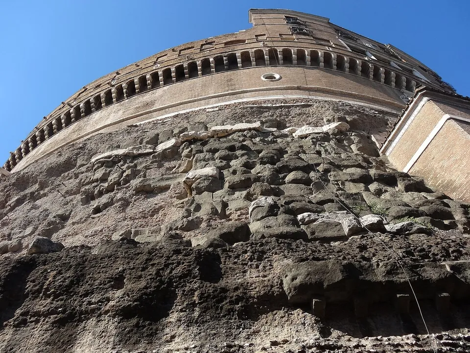 Looking up the curved wall of the drum, with rough, weathered stone blocks in the lower part, smoother masonry above, and a ring of small openings beneath a parapet at the top against a blue sky.