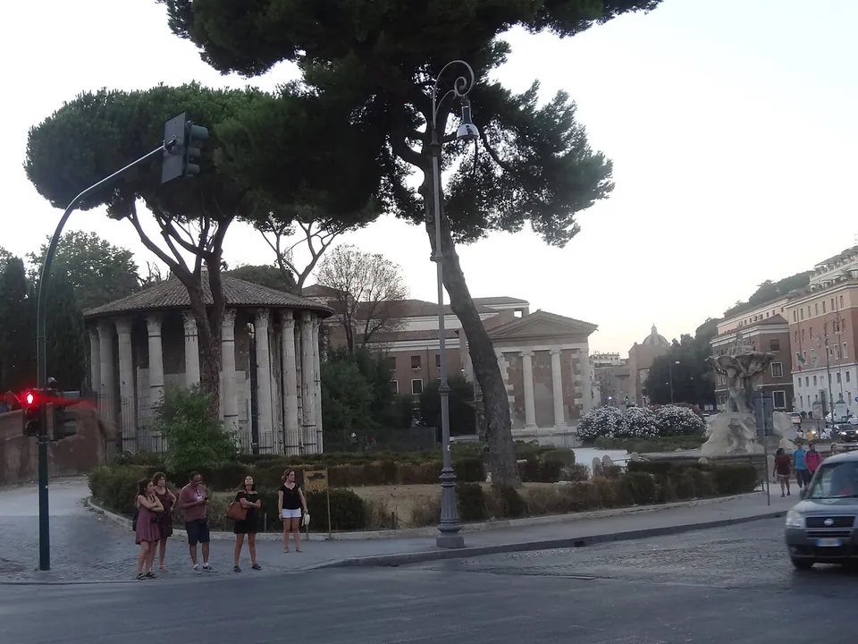 A street corner at dusk with a traffic light and pedestrians, umbrella pines above a hedged garden, a round colonnaded temple among the trees at left and a rectangular temple with a pediment behind, and a fountain with flowers at right.