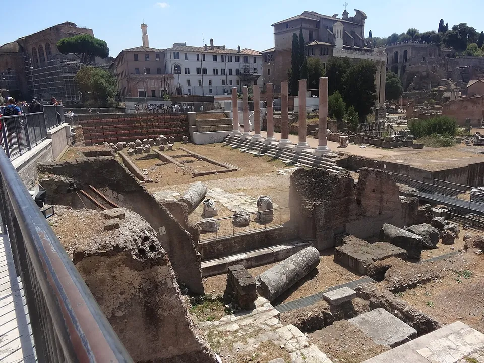 Excavated ground seen from a railed walkway, with fallen column drums and low walls in the foreground and a row of seven pinkish columns standing on stepped bases beyond, with modern buildings and trees behind.