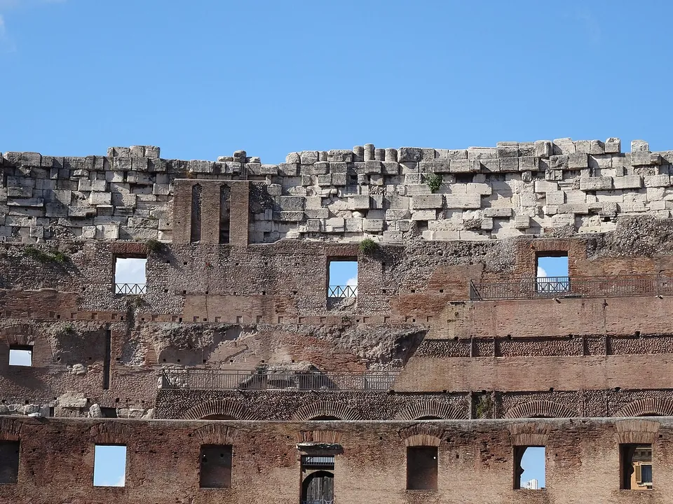 The jagged crest of a ruined wall against a blue sky, a course of large pale stone blocks above storeys of brick wall pierced by small rectangular openings, some fitted with modern railings.