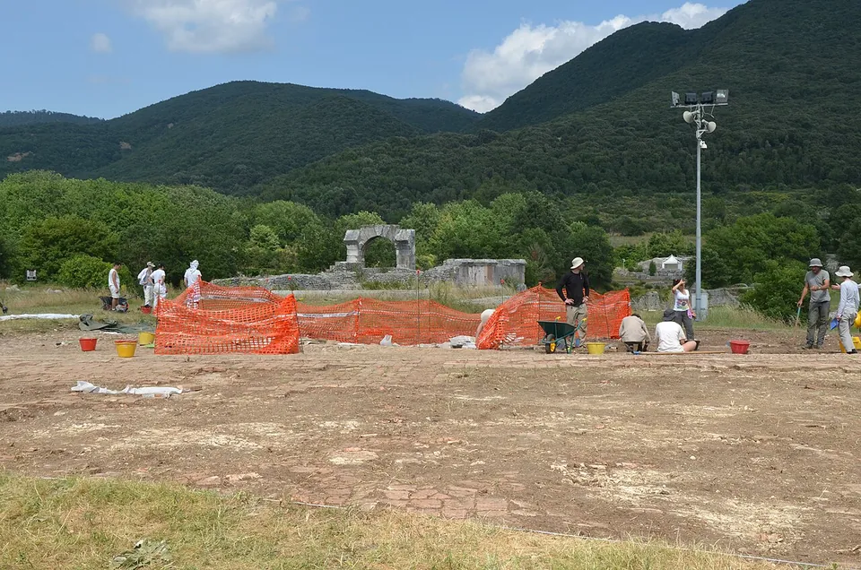A broad flat area of bare excavated ground with orange safety netting, buckets and a wheelbarrow, archaeologists at work, a single stone arch standing at the far edge, and steep wooded hills behind.