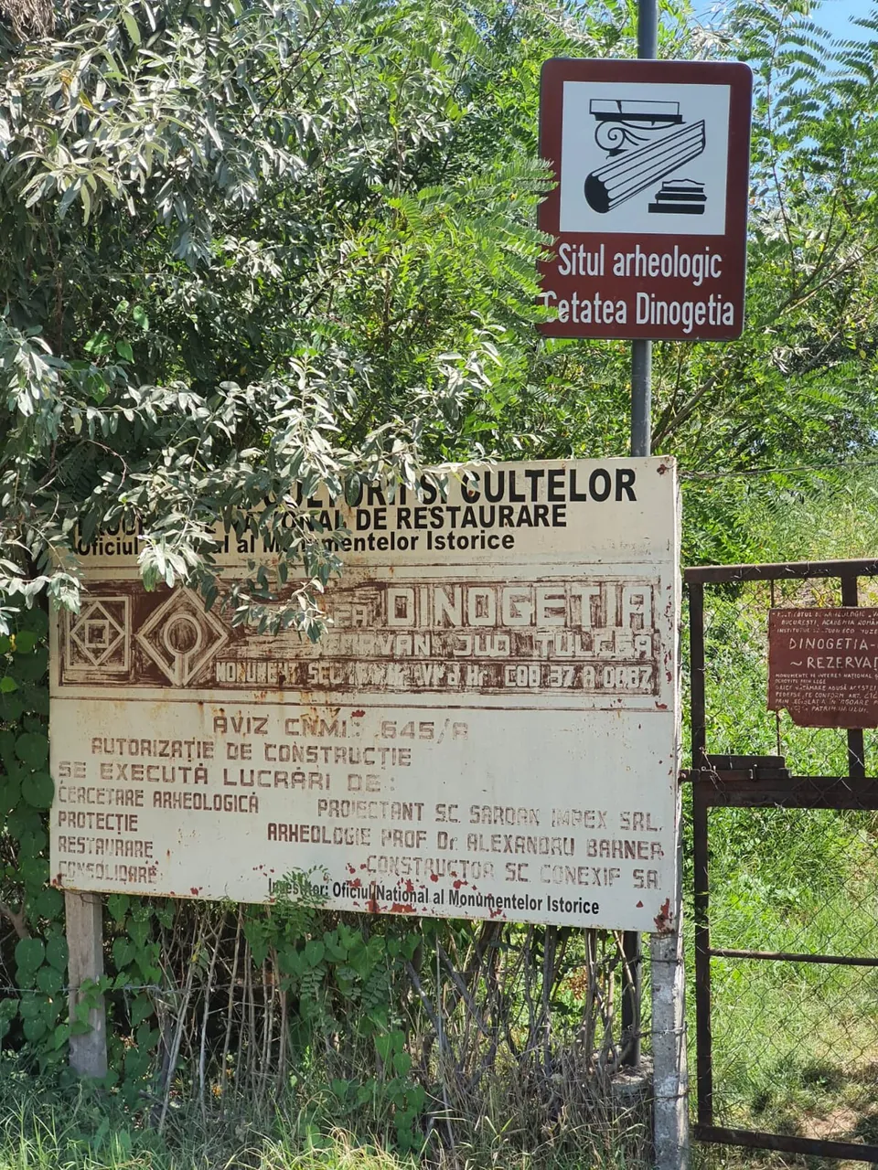 A brown heritage sign reading "Situl arheologic Cetatea Dinogetia" above an old rusted notice board about restoration works, beside a metal gate in a fence overgrown with trees.