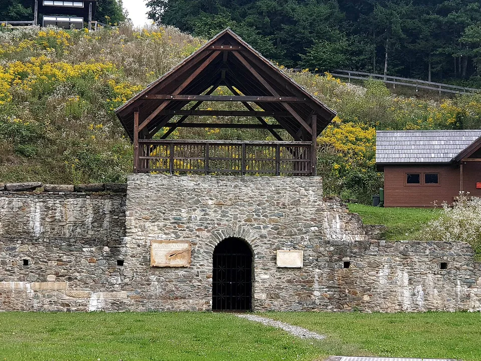 A stretch of stone wall with an arched doorway at its centre, topped by an open timber-roofed platform, standing at the edge of a grassy arena.