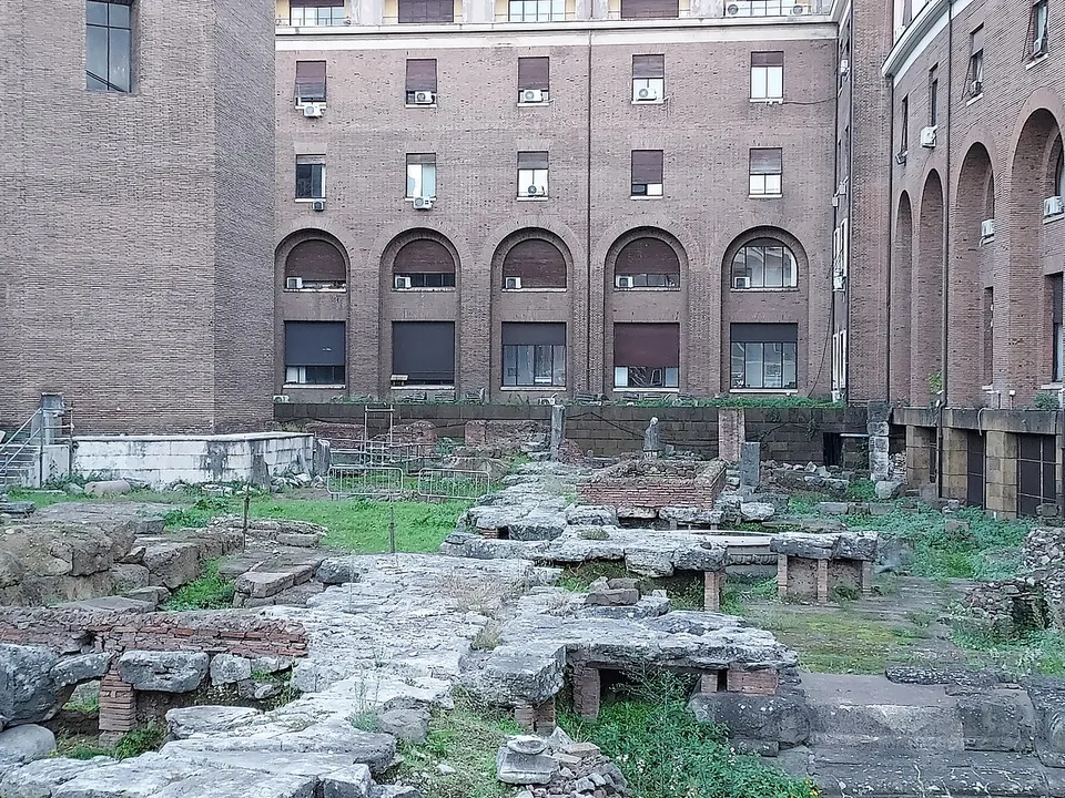 Seen from the street, broad grey stone slabs, some propped on short brick posts, spread across a grassy excavation in front of a tall brick building with arched windows.