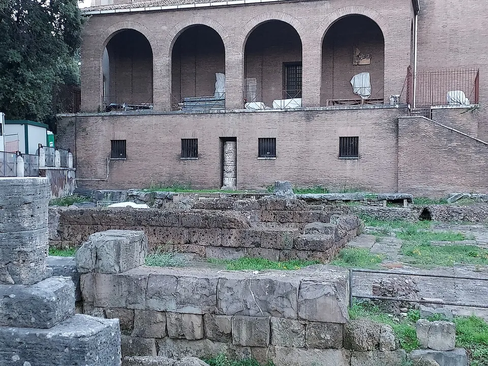 Walls of large squared stone blocks in the foreground, and behind them the brick flank of the church with a row of tall open arches along its upper storey.