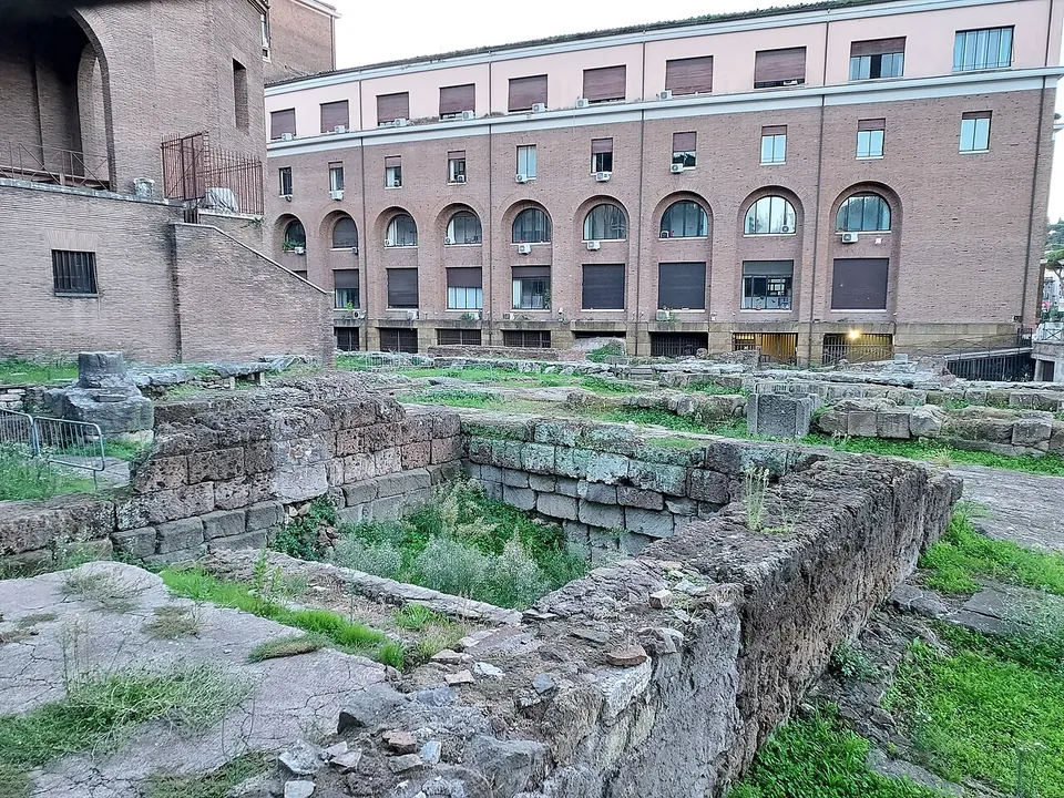 Close view of low walls of squared grey-brown tufa blocks laid in courses, worn at the edges and patched with grass, in front of a modern building.