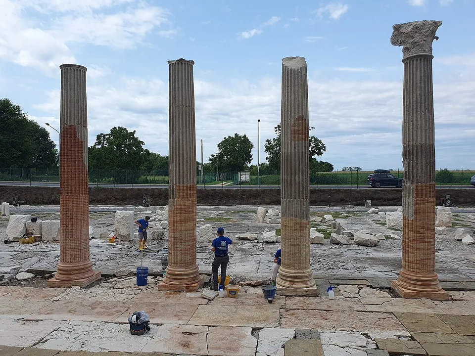 Four columns, patched with red brick and one topped by a carved capital, standing on a paving of large pale stone slabs, while archaeologists work among scattered blocks behind them.