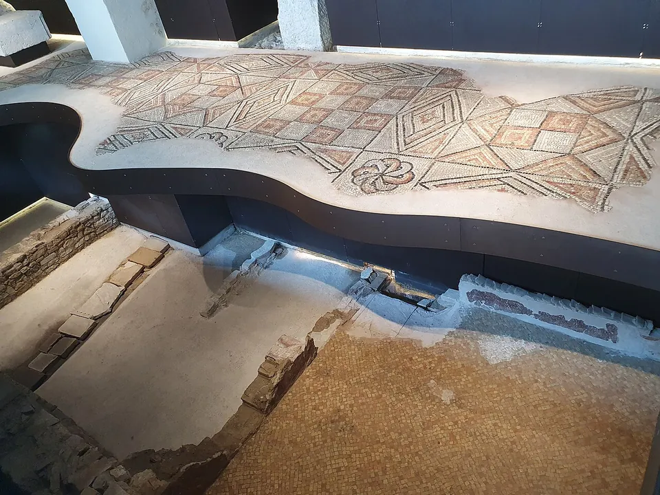 A mosaic floor of lozenges and squares laid on a curved raised platform indoors, with lower walls, a stone threshold and a plain brick-coloured floor visible in the excavated level beneath it.