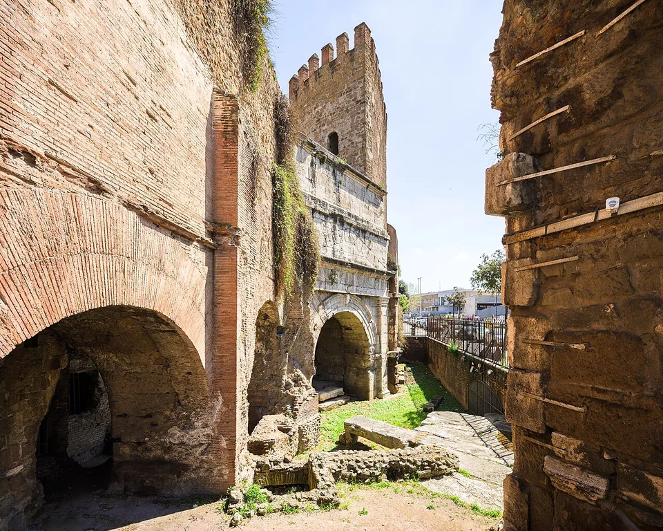 A narrow passage between two high brick walls with round-arched openings, leading to a gate of pale stone blocks with an arched opening, a crenellated tower rising above it and a strip of grass and paving in front.